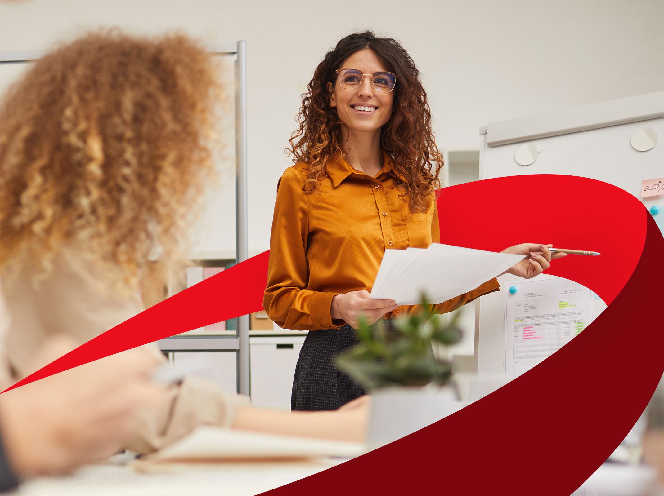 A business professional communicating with others in front of a white board.  She is hold some papers and in smiling in a business setting.