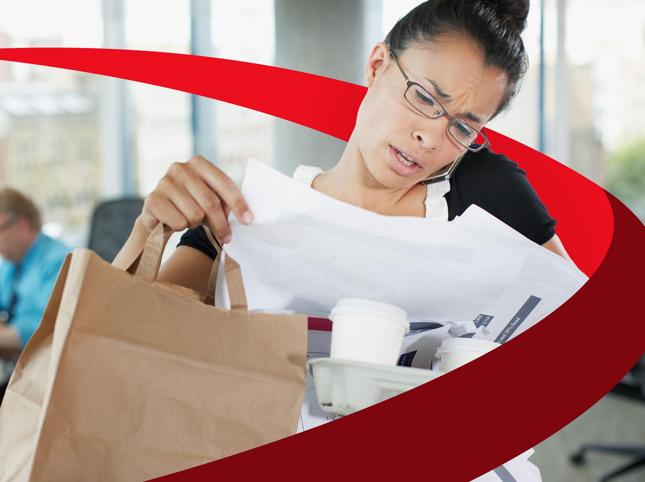 A stressed business professional balancing papers, a phone, coffee cups and a bag.  She is in an office setting.